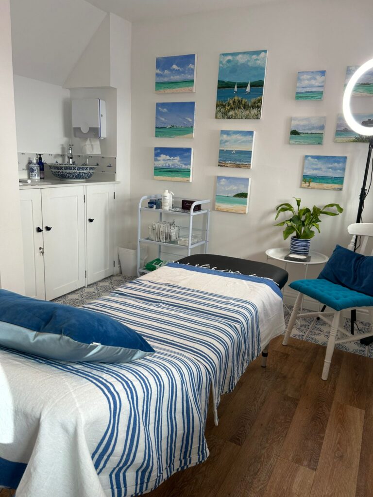 A calm, softly lit treatment room at Blue Bird Aesthetics featuring a neatly made treatment bed with a blue‑striped blanket, ocean artwork on the wall, and a clean clinical sink area, creating a serene and professional environment.