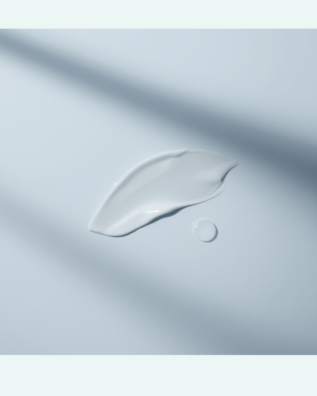 A minimalist flat‑lay showing a soft smear of white moisturiser and a single serum droplet on a pale blue‑grey background